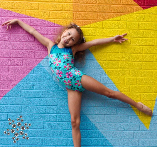 A gymnast in front of a colorful wall in a teal gymnastics leotard.